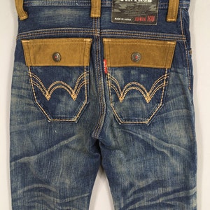 May include: Blue denim jeans with brown suede-like pocket accents and decorative stitching. The jeans feature a faded wash and a branded tag that reads "EXCLUSIVE VINTAGE" and "EDWIN XU".