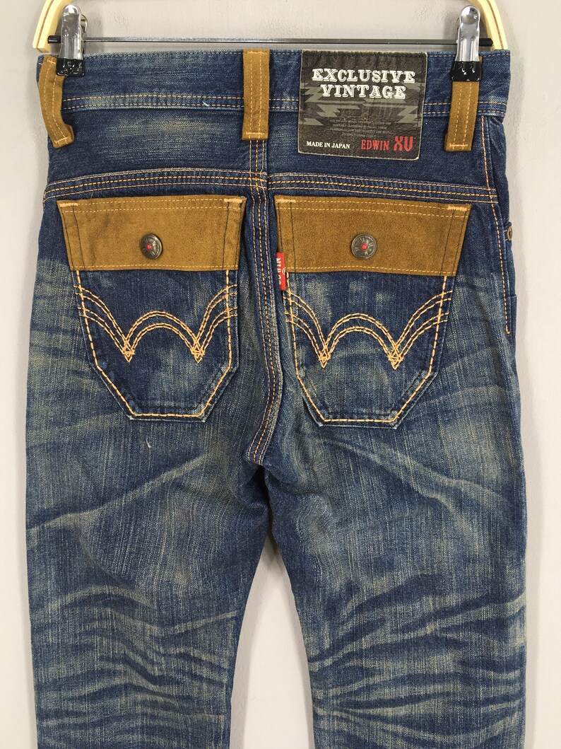 May include: Blue denim jeans with brown suede-like pocket accents and decorative stitching. The jeans feature a faded wash and a branded tag that reads "EXCLUSIVE VINTAGE" and "EDWIN XU".