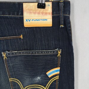 May include: Close-up of dark blue denim jeans with yellow stitching. The jeans feature a back pocket with a distinctive yellow stitched design and a small label with the text "XV-FUNCTION". The jeans are hanging on a hanger.