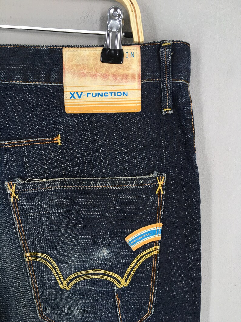 May include: Close-up of dark blue denim jeans with yellow stitching. The jeans feature a back pocket with a distinctive yellow stitched design and a small label with the text "XV-FUNCTION". The jeans are hanging on a hanger.