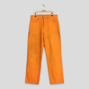 May include: Bright orange denim pants hanging on a gold hanger. The pants have a classic five-pocket design with a button and zipper closure. There are some stains on the front of the pants. The pants are a solid color.