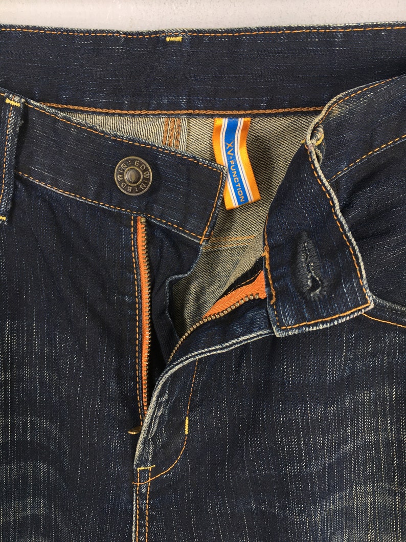 May include: Close-up of dark blue denim jeans with orange stitching and a visible zipper. The jeans have a button closure with the text "WIN ED BD" and a tag that reads "XV-FUNCTION" in white and blue.