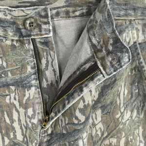 May include: Close-up of camouflage pants with an open zipper and button closure. The pants feature a pattern of green, brown, and gray, resembling a forest or woodland environment. The zipper is gold-colored.