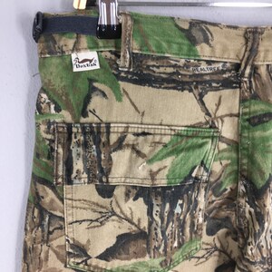 Puede incluir: Primer plano de pantalones de camuflaje con un logotipo de Realtree y una etiqueta Duxbak. Los pantalones presentan un patr&oacute;n de bosque en tonos verdes, marrones y beige. El bolsillo trasero es visible y las trabillas est&aacute;n en la parte superior.