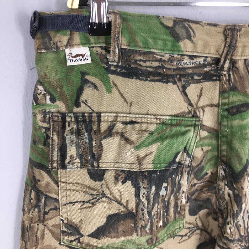 Puede incluir: Primer plano de pantalones de camuflaje con un logotipo de Realtree y una etiqueta Duxbak. Los pantalones presentan un patr&oacute;n de bosque en tonos verdes, marrones y beige. El bolsillo trasero es visible y las trabillas est&aacute;n en la parte superior.