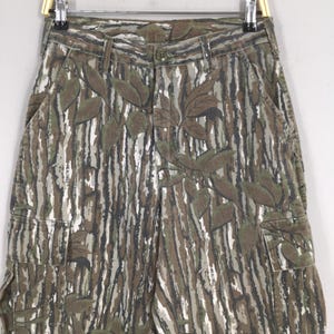 May include: Camouflage cargo pants with a brown, green, and white pattern. The pants feature a leaf design and multiple pockets. The pants are hanging on a metal hanger.