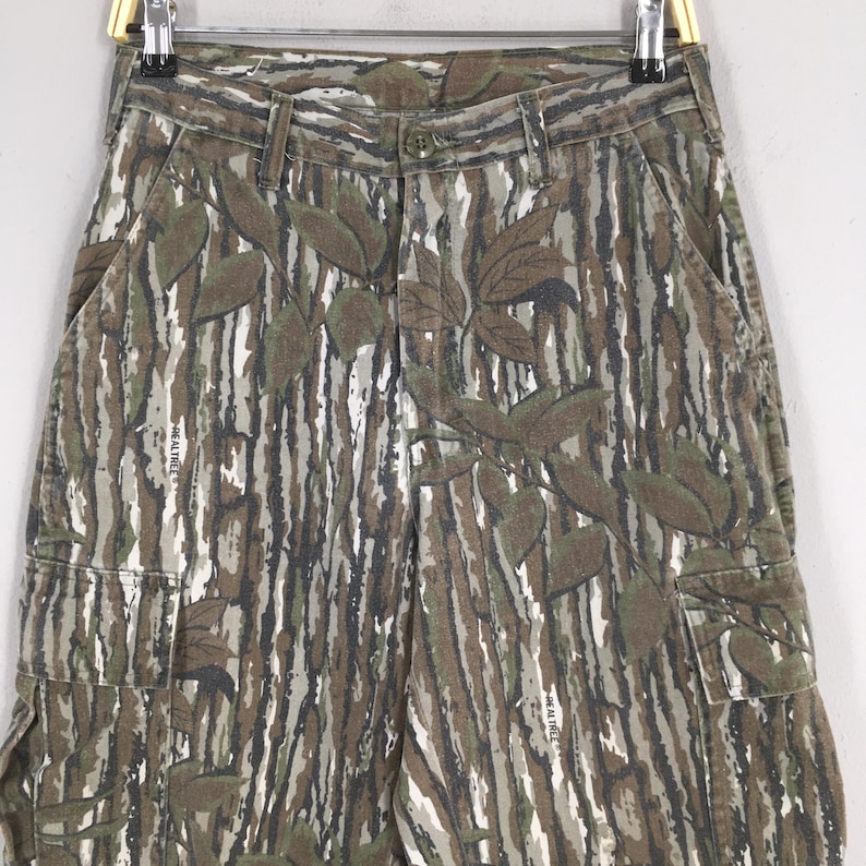 May include: Camouflage cargo pants with a brown, green, and white pattern. The pants feature a leaf design and multiple pockets. The pants are hanging on a metal hanger.