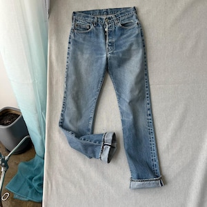 Könnte beinhalten: Hellblaue Jeans aus Denim mit Knopfleiste, fünf Taschen und geradem Bein. Der Stoff hat eine verwaschene Optik mit sichtbaren Tragespuren an den Hüften und hochgekrempelten Beinabschlüssen, die eine Webkante sichtbar machen.