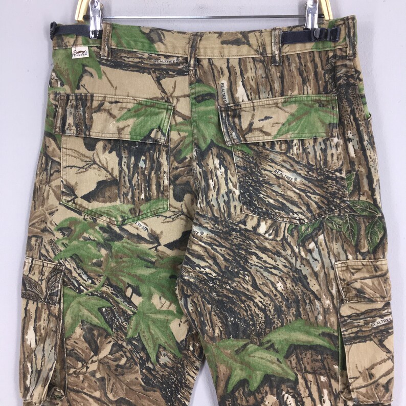 Puede incluir: Pantalones cargo de camuflaje con m&uacute;ltiples bolsillos y un patr&oacute;n Realtree. Los pantalones presentan una mezcla de colores marr&oacute;n, verde y beige, dise&ntilde;ados para integrarse en entornos naturales. Los pantalones est&aacute;n colgados de una percha.