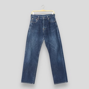 Puede incluir: Vaqueros de mezclilla azul oscuro colgados de una percha dorada. Los vaqueros tienen bragueta con botones, diseño clásico de cinco bolsillos y remaches de cobre. La mezclilla tiene un aspecto descolorido y desgastado.