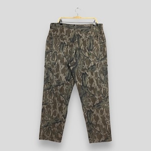 May include: Camouflage cargo pants with multiple pockets, hanging on a gold hanger. The pants feature a brown, green, and tan pattern. The pants have a button and zipper closure and belt loops.