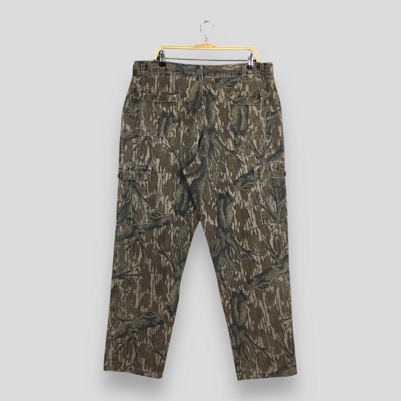 May include: Camouflage cargo pants with multiple pockets, hanging on a gold hanger. The pants feature a brown, green, and tan pattern. The pants have a button and zipper closure and belt loops.