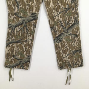 Pu&ograve; includere: Primo piano di pantaloni mimetici con un motivo dettagliato di rami di alberi e fogliame nelle tonalit&agrave; del marrone, verde e beige. I pantaloni hanno una tasca cargo e coulisse sull'orlo inferiore.