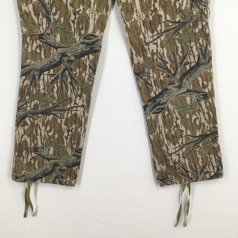 Pu&ograve; includere: Primo piano di pantaloni mimetici con un motivo dettagliato di rami di alberi e fogliame nelle tonalit&agrave; del marrone, verde e beige. I pantaloni hanno una tasca cargo e coulisse sull'orlo inferiore.