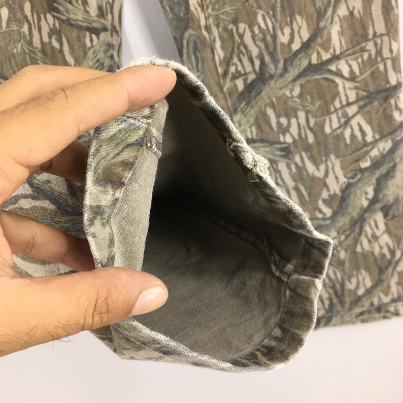 May include: Close-up of camouflage pants, featuring a detailed pattern of brown, tan, and green resembling a forest. The fabric is visible, with the inside of the pant leg showing a lighter shade of the same color scheme.