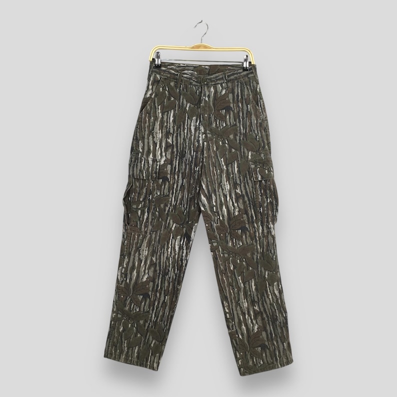 May include: Green and gray camouflage cargo pants with multiple pockets. The pants are hanging on a gold-colored hanger. The camouflage pattern has a vertical stripe design.