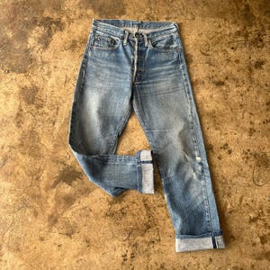 Puede incluir: Vaqueros de mezclilla azul claro con bragueta de botones y un lavado descolorido. Los vaqueros tienen un diseño clásico de cinco bolsillos y están doblados en los tobillos. La mezclilla tiene algo de desgaste, dándole un aspecto vintage.