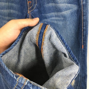 May include: Close-up of blue denim jeans with orange stitching details. The jeans show signs of wear and fading, with a distressed look. The inside of the jeans' hem is visible, showcasing the fabric's texture and construction.