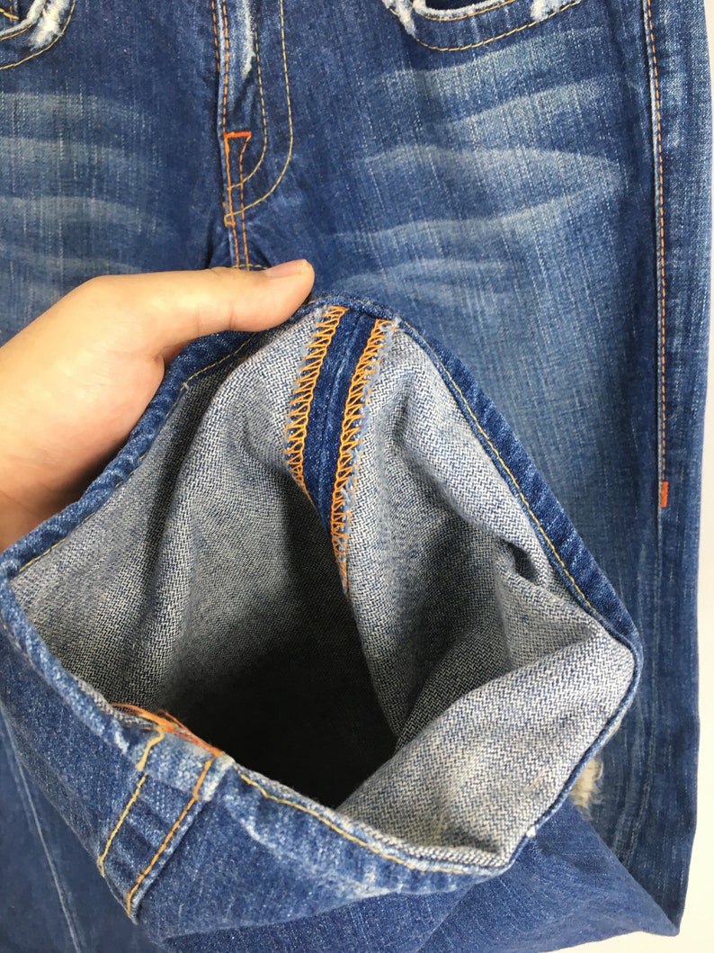 May include: Close-up of blue denim jeans with orange stitching details. The jeans show signs of wear and fading, with a distressed look. The inside of the jeans' hem is visible, showcasing the fabric's texture and construction.