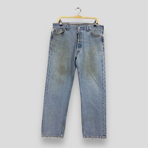 May include: Light blue denim jeans with a button fly, hanging on a gold-colored hanger. The jeans show signs of wear, with faded areas and a small tear on the right leg. A classic, casual style.