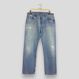 May include: Light blue denim jeans with distressed details, including rips and fading. The jeans are hanging on a gold-colored hanger. The jeans have a classic five-pocket design and a button fly.