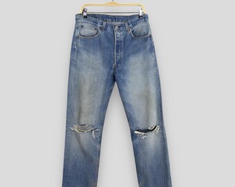May include: Light blue denim jeans with distressed details, including ripped knees. The jeans are hanging on a gold-colored hanger. The jeans have a button fly and classic five-pocket design.