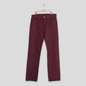 May include: Burgundy-colored denim jeans hanging on a wooden hanger. The jeans have a classic five-pocket design and a straight leg cut. The fabric appears to be slightly faded, giving them a vintage look. The jeans are displayed against a light gray background.