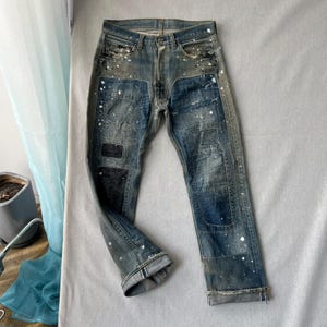Könnte beinhalten: Blaue Jeans aus Denim mit verwaschener Optik, weißen Farbspritzern und mehreren rechteckigen Flicken in verschiedenen Blau- und Schwarztönen. Die Jeans hat einen Knopfverschluss, fünf Taschen und ausgefranste, umgeschlagene Beinabschlüsse.