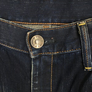 May include: Close-up of dark blue denim jeans. The jeans feature a silver button with the text "EDWIN & CO" and orange stitching. The jeans have a classic five-pocket design and a button and zipper closure.