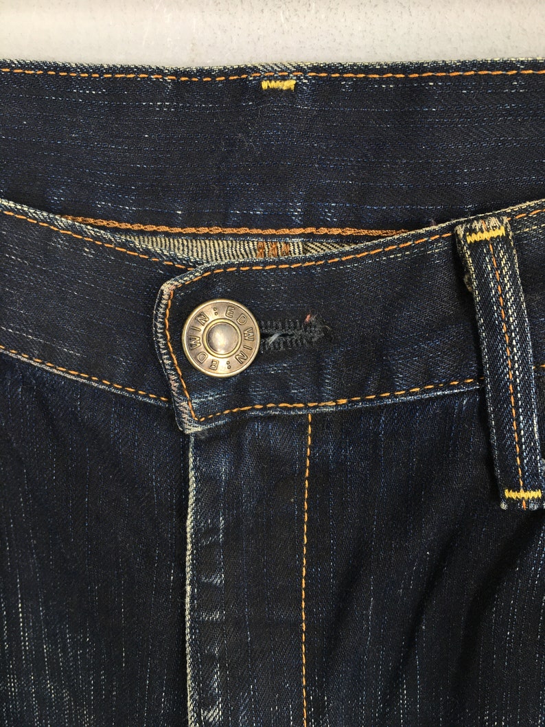May include: Close-up of dark blue denim jeans. The jeans feature a silver button with the text "EDWIN & CO" and orange stitching. The jeans have a classic five-pocket design and a button and zipper closure.