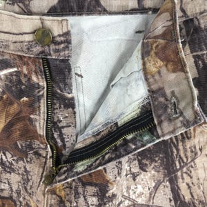 K&ouml;nnte beinhalten: Nahaufnahme einer Camouflage-Hose mit einem teilweise ge&ouml;ffneten schwarzen Rei&szlig;verschluss. Die Hose hat einen Knopfverschluss und ein realistisches Waldmuster in Braun-, Gr&uuml;n- und Beiget&ouml;nen. Der Stoff scheint strapazierf&auml;hig zu sein.
