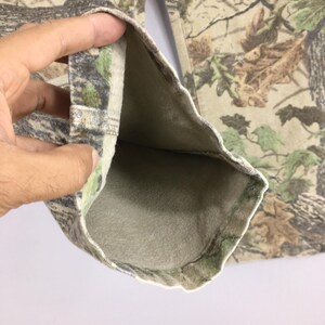 May include: Close-up of camouflage pants, featuring a detailed pattern of brown, green, and tan foliage. The interior lining is a solid, light beige color. The fabric is folded, revealing the inside of the pants.