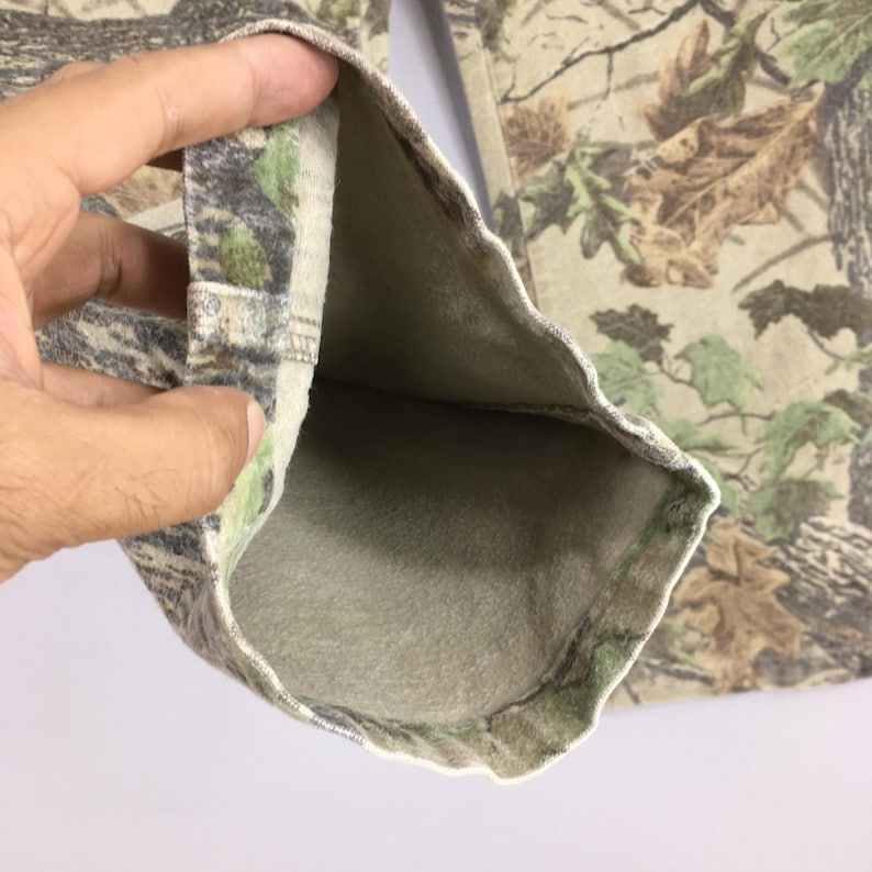 May include: Close-up of camouflage pants, featuring a detailed pattern of brown, green, and tan foliage. The interior lining is a solid, light beige color. The fabric is folded, revealing the inside of the pants.