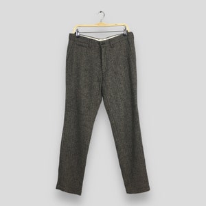 Può includere: Pantaloni marroni a spina di pesce appesi a una gruccia di legno. I pantaloni hanno una chiusura con bottone e cerniera, con tasche. Il tessuto è un misto lana strutturato. La vita misura circa 81 cm.