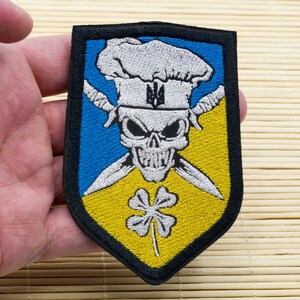 Può includere: Toppa ricamata con un teschio che indossa un cappello da cuoco con un tridente ucraino sul cappello. Il teschio tiene due coltelli incrociati. La toppa è su uno sfondo giallo e blu con un quadrifoglio.