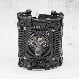 Puede incluir: Pulsera de cuero negro con un diseño de cabeza de lobo plateado. La pulsera está trenzada y tiene tachuelas metálicas.