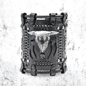 May include: Black leather bracelet with a silver skull design and metal accents. The bracelet has a braided leather strap and is decorated with studs.