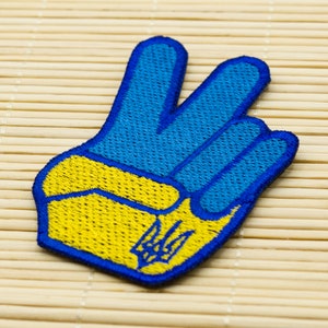 Può includere: Una toppa a forma di segno di pace blu e gialla con il simbolo del tridente ucraino al centro.