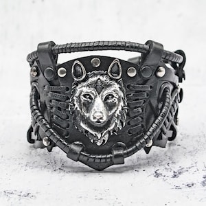 Può includere: Bracciale a polsino in pelle nera con una testa di lupo argentata dettagliata. Il bracciale ha una parte superiore intrecciata ed è adornato con borchie argentate. Il design incorpora elementi in pelle cuciti e rivettati.