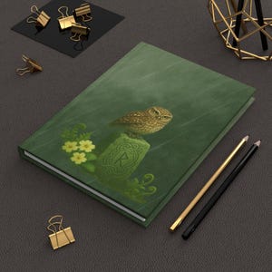 May include: A green journal featuring an illustration of a small owl perched on a stone with Celtic knotwork. Yellow flowers and fern fronds are also depicted. Gold and black pencils and gold binder clips are also visible.