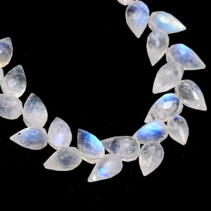 Pode incluir: Um colar de contas facetadas de pedra da lua em forma de pêra. As contas são claras com flashes iridescentes azuis e brancos. As contas são enfiadas em um fio prateado, em um fundo preto.