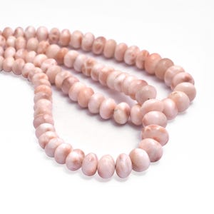 Puede incluir: Una hebra de cuentas de concha rosa y blanca. Las cuentas son de forma ovalada y tienen un acabado liso y pulido.