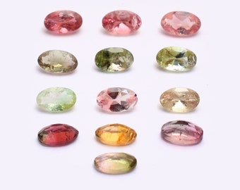 May include: Assortment of oval-shaped gemstones in various colors, including pink, green, yellow, and clear. The gemstones are arranged in rows on a white surface.