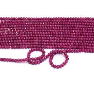 May include: Strands of faceted, deep red ruby beads. The beads are strung together, forming a dense, textured surface. A few strands are arranged in loops, showcasing the color and shape of the gemstones. The background is plain white.