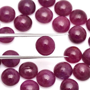 May include: A selection of polished, round, deep red gemstones. One gem is held by tweezers, displaying its smooth surface and rich colour. The gems are scattered on a white surface, highlighting their vibrant hue. These are likely rubies.