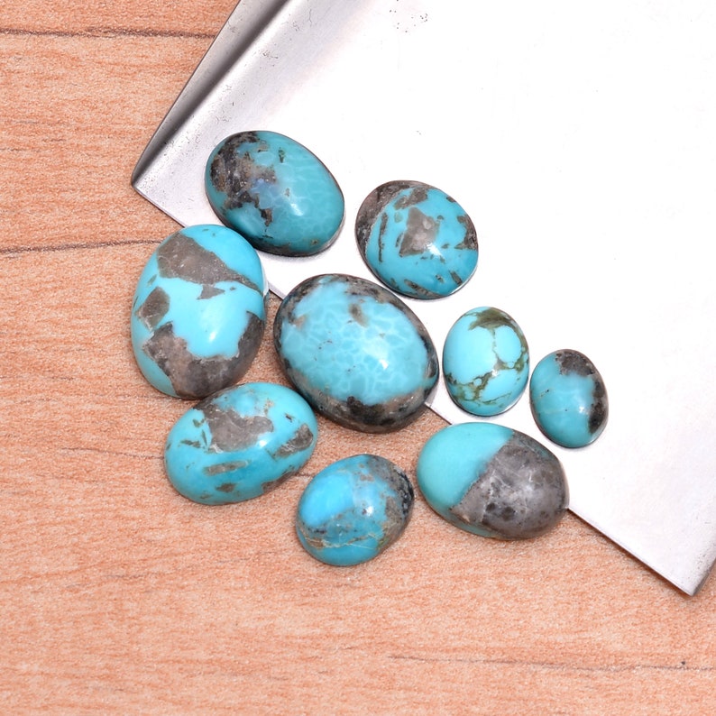 May include: A collection of nine oval-shaped turquoise cabochons with a blue-green color and black matrix. The stones are arranged on a silver surface.