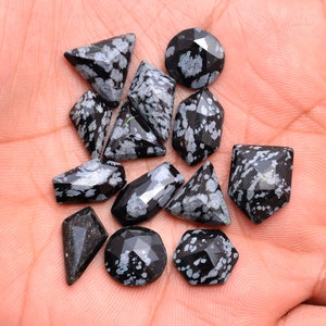 May include: A collection of twelve snowflake obsidian gemstone cabochons in various shapes and sizes. The stones are black with white flecks.