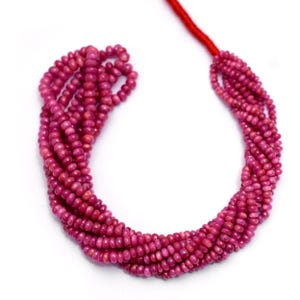 May include: Strands of small, round, deep pink gemstones, likely rubies, are displayed against a white background. The gemstones are strung together on a red cord, creating a necklace or bead strand. The beads have a polished appearance.