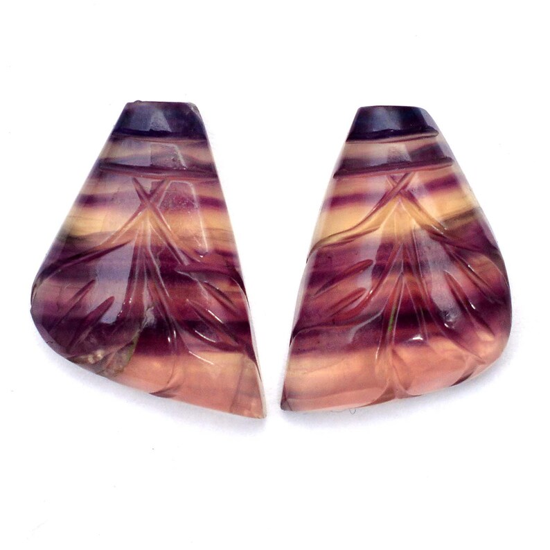 K&ouml;nnte beinhalten: Zwei geschnitzte Fluorit-Cabochons mit einem lila, rosa und gelben Farbmuster. Die Cabochons sind tropfenf&ouml;rmig und haben eine glatte, polierte Oberfl&auml;che.