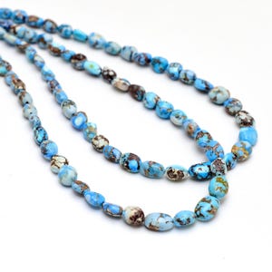 Könnte beinhalten: Stränge türkisfarbener Edelsteinperlen. Die oval geformten Perlen weisen eine Mischung aus blauen, braunen und weißen Farbtönen auf. Die Perlen sind aufgefädelt und bilden eine Halskette oder ein Armband. Der Hintergrund ist schlicht weiß.